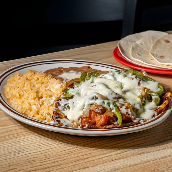 A Mexican dish with melted cheese, peppers, onions, rice, and refried beans, with tortillas in the background.